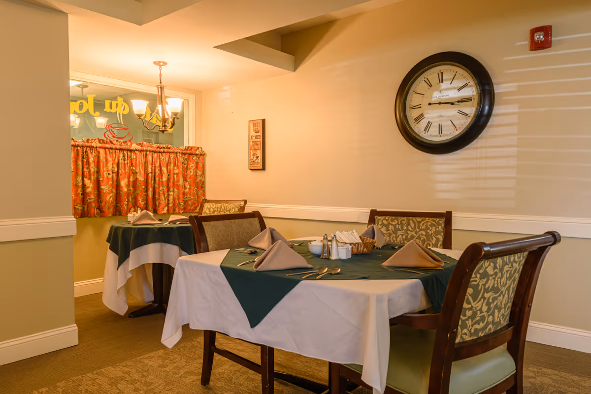 A cozy dining area with two tables covered in white and green tablecloths, each set with folded napkins, silverware, and condiments. The room features a large wall clock with Roman numerals and a window with floral curtains. Warm lighting creates a welcoming atmosphere.