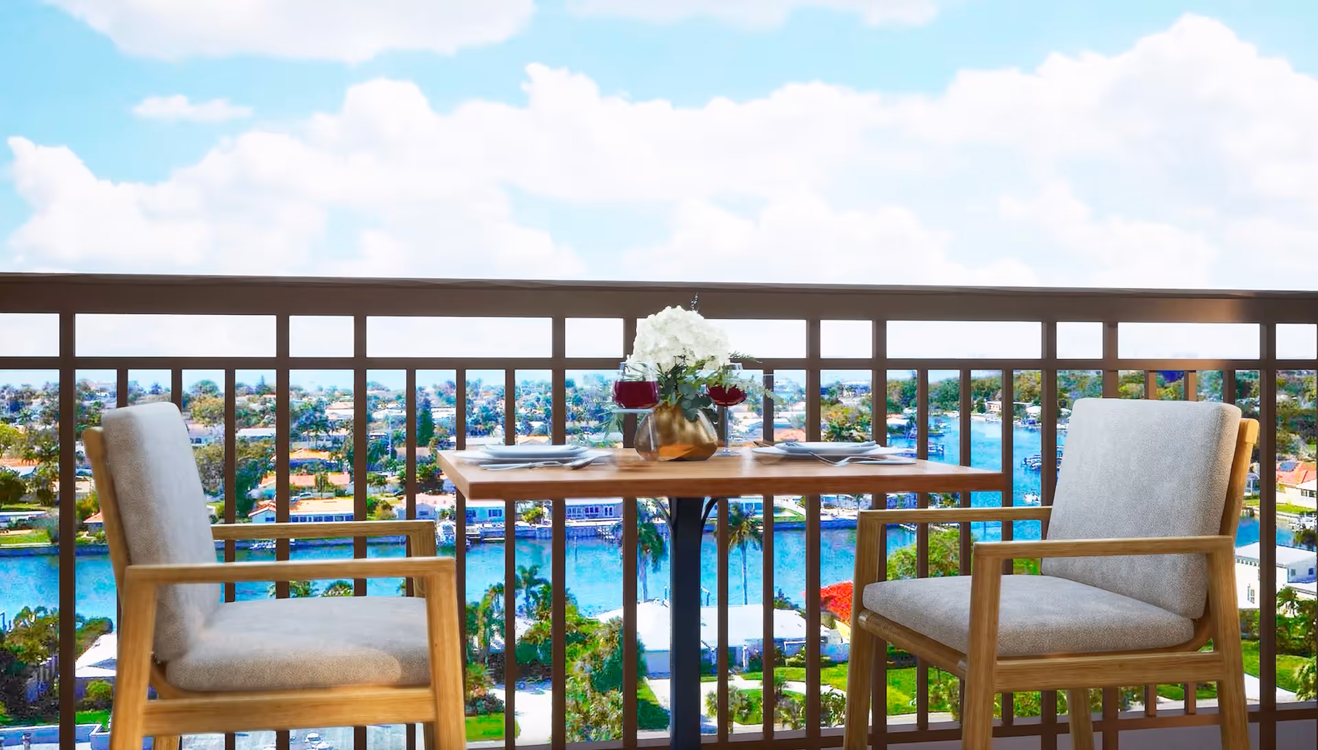 A balcony with a wooden table set for two, featuring two cushioned wooden chairs and a small vase with white flowers. The balcony overlooks a scenic waterfront neighborhood with houses, trees, and a bright blue sky with scattered clouds.