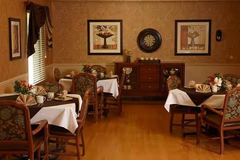 A warmly lit senior dining room with white tablecloths, round tables, and wall art.