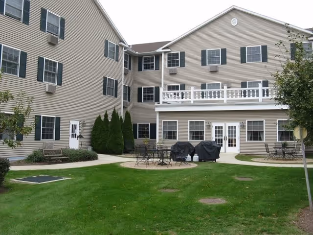 Outdoor courtyard area of a senior living facility with green grass, several round patio tables with chairs, two covered grills, and a beige multi-story building with multiple windows and a balcony in the background.