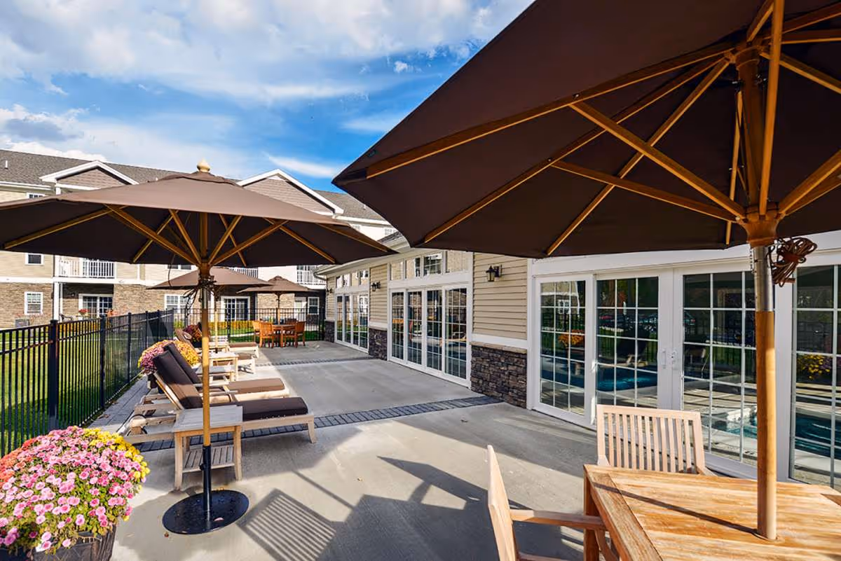Outdoor patio area at Summit at Glenwyck featuring several lounge chairs with cushions under large brown umbrellas, a wooden table with chairs, potted flowers, and a building with large glass doors and windows in the background under a partly cloudy sky.