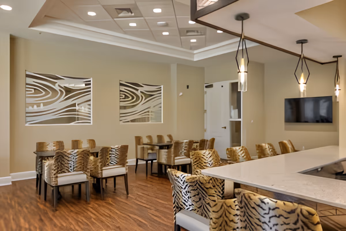 Interior view of a dining area in a senior living facility featuring multiple tables with chairs upholstered in a tiger stripe pattern. The room has wooden flooring, beige walls with decorative glass panels, pendant lights hanging from the ceiling, and a wall-mounted flat screen TV.
