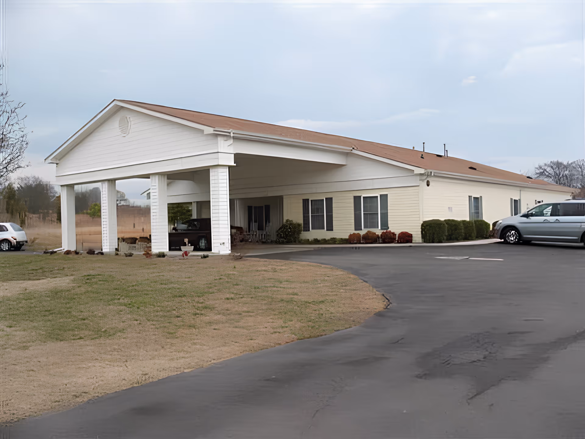 Exterior view of a single-story building with a covered entrance supported by white columns, a brown roof, and several windows. There is a paved driveway curving in front of the building, with some parked vehicles visible on the right side. The surrounding area has grass and some small bushes near the building.