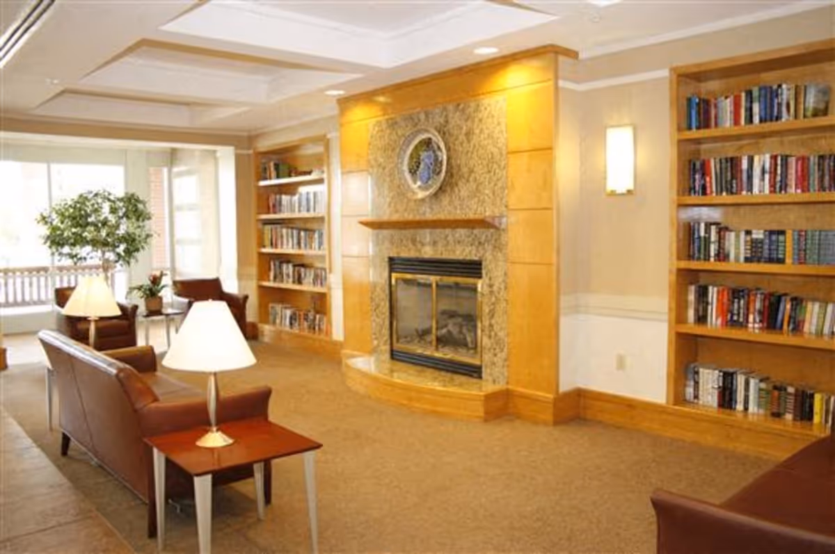 A cozy living room area with a fireplace surrounded by a stone and wood mantel. There are built-in wooden bookshelves filled with books on either side of the fireplace. The room has brown leather chairs and sofas, small wooden side tables with lamps, and a large window letting in natural light. A potted plant is visible near the window.