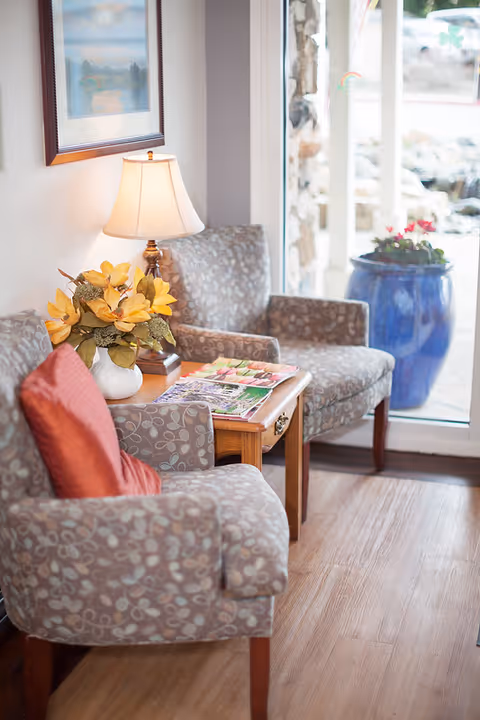 A cozy seating area with two patterned armchairs, one with a red cushion, and a wooden side table between them holding a lamp, a vase with yellow flowers, and magazines. A large window shows a blue planter with flowers outside.