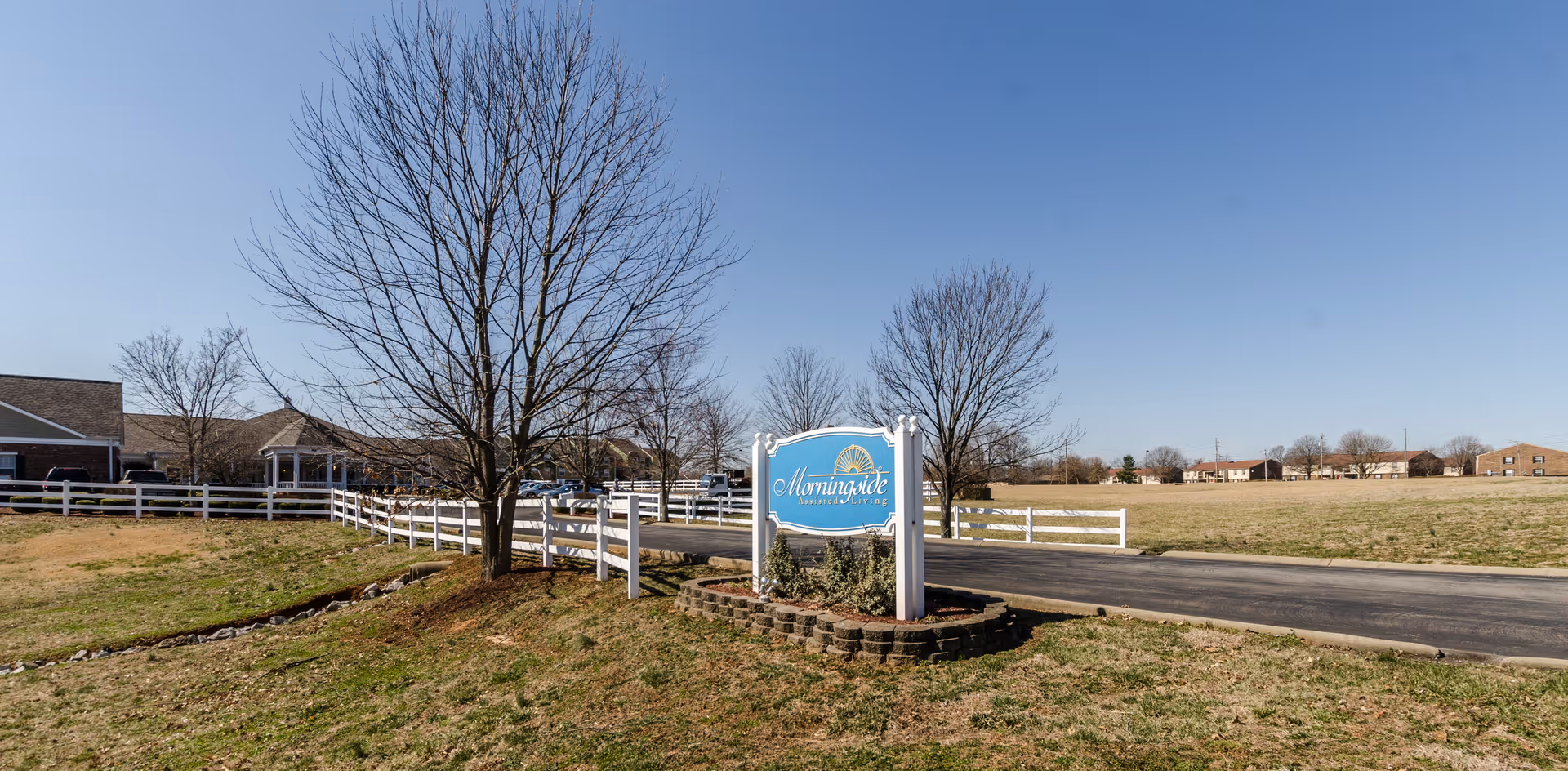 Entrance sign and driveway of a senior living community with a white fence, leafless trees, and buildings in the background under a clear blue sky.