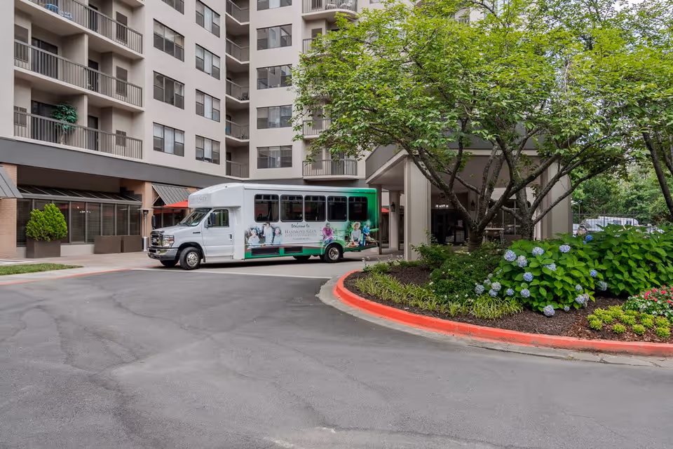 Exterior view of Hammond Glen Retirement Community showing a multi-story building with balconies, a white shuttle bus parked near the entrance, and landscaped greenery including trees and flowering plants.