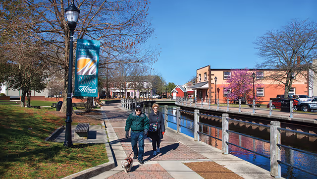 A man and woman walking a small dog along a paved pathway next to a canal with railings. There are buildings, trees, and parked cars visible under a clear blue sky. A street lamp with a banner that reads 'Milford' is also visible.