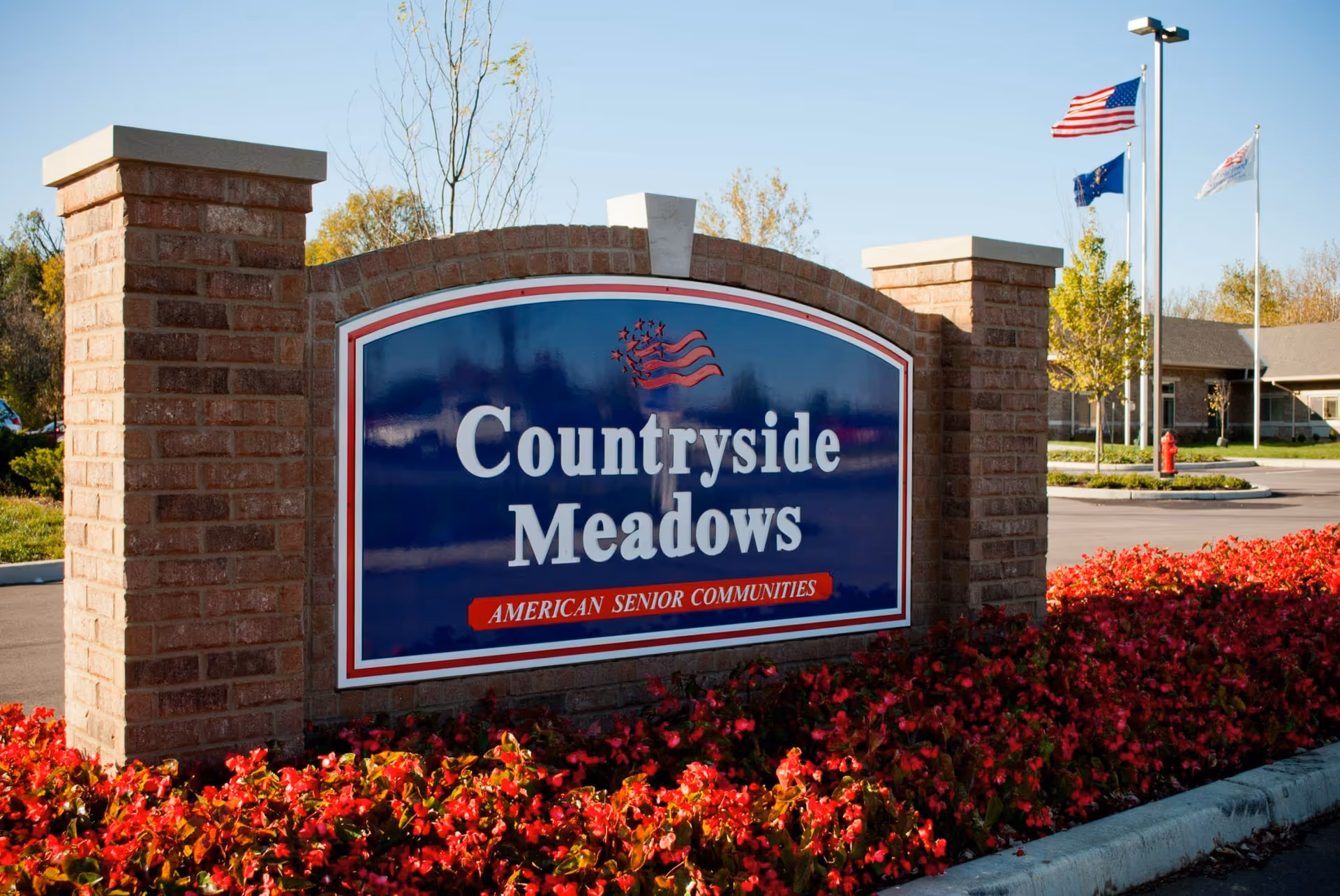 A large brick sign at the entrance of Countryside Meadows, an American senior community, surrounded by red flowers with a building and three flagpoles flying the American flag and other flags in the background under a clear blue sky.
