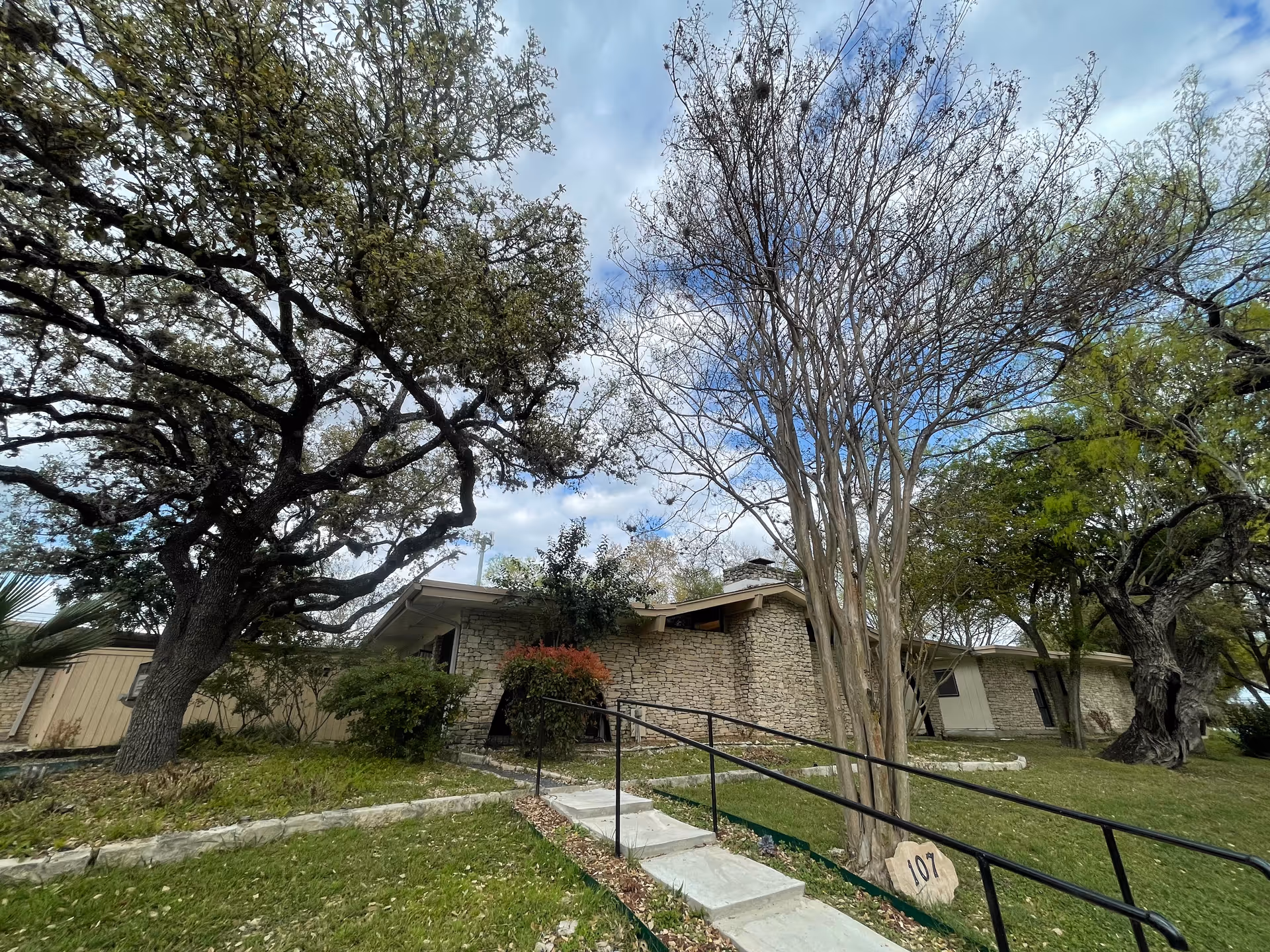 Exterior view of a single-story stone building surrounded by trees and greenery under a partly cloudy sky. A concrete pathway with a black handrail leads up to the entrance, and a stone marker with the number 107 is visible near the pathway.
