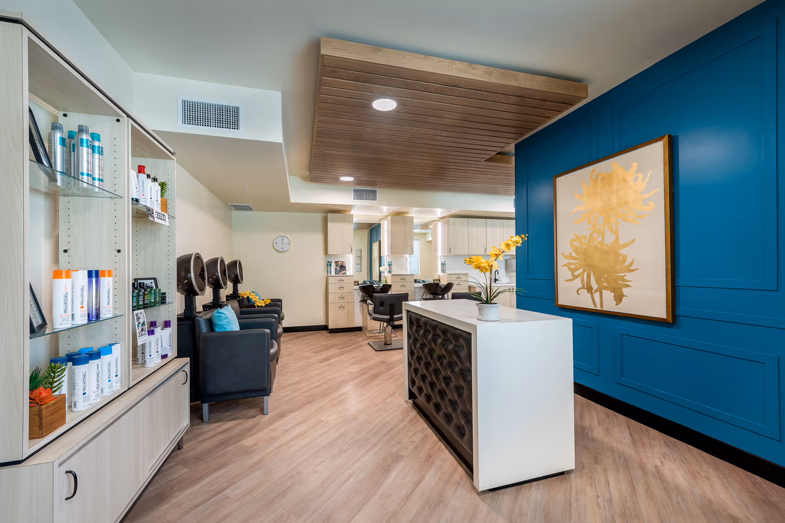 Interior view of a modern salon area with a blue accent wall featuring a large gold floral artwork. There is a white reception desk with a tufted black front panel and a potted yellow orchid on top. To the left, there is a display cabinet with various hair care products. In the background, there are black salon chairs with hair dryers and a clock on the wall.