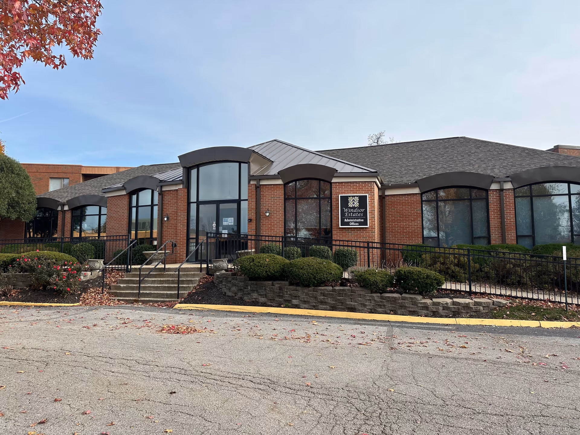 Exterior view of a single-story brick building with large windows and a central entrance with steps and a ramp. The building has a sign that reads 'Windsor Estates Administrative Offices'. There are bushes and a tree with red leaves in front of the building, and fallen leaves scattered on the ground.