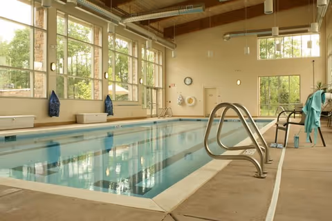 Indoor swimming pool area with large windows letting in natural light, metal handrails for pool entry, chairs and towels on the side, and a clock on the far wall.