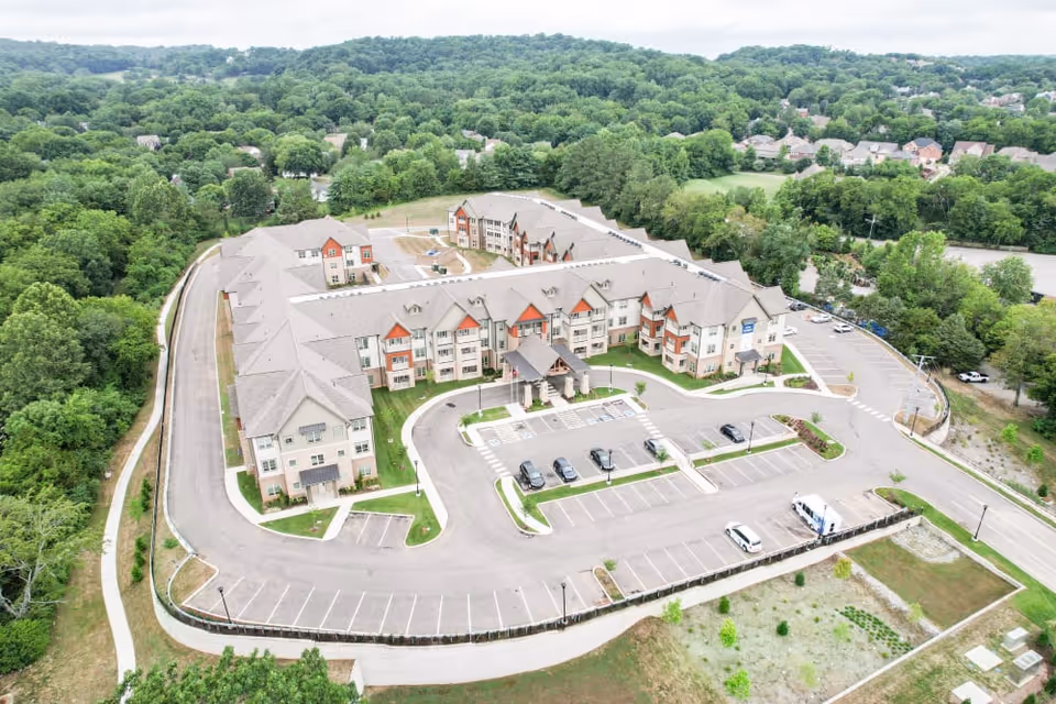 Aerial view of a large senior living facility named Harmony At Bellevue, surrounded by trees and greenery. The building is three stories tall with a beige and red exterior, featuring a covered entrance and a spacious parking lot with several cars parked. The facility is situated in a suburban area with residential neighborhoods visible in the background.