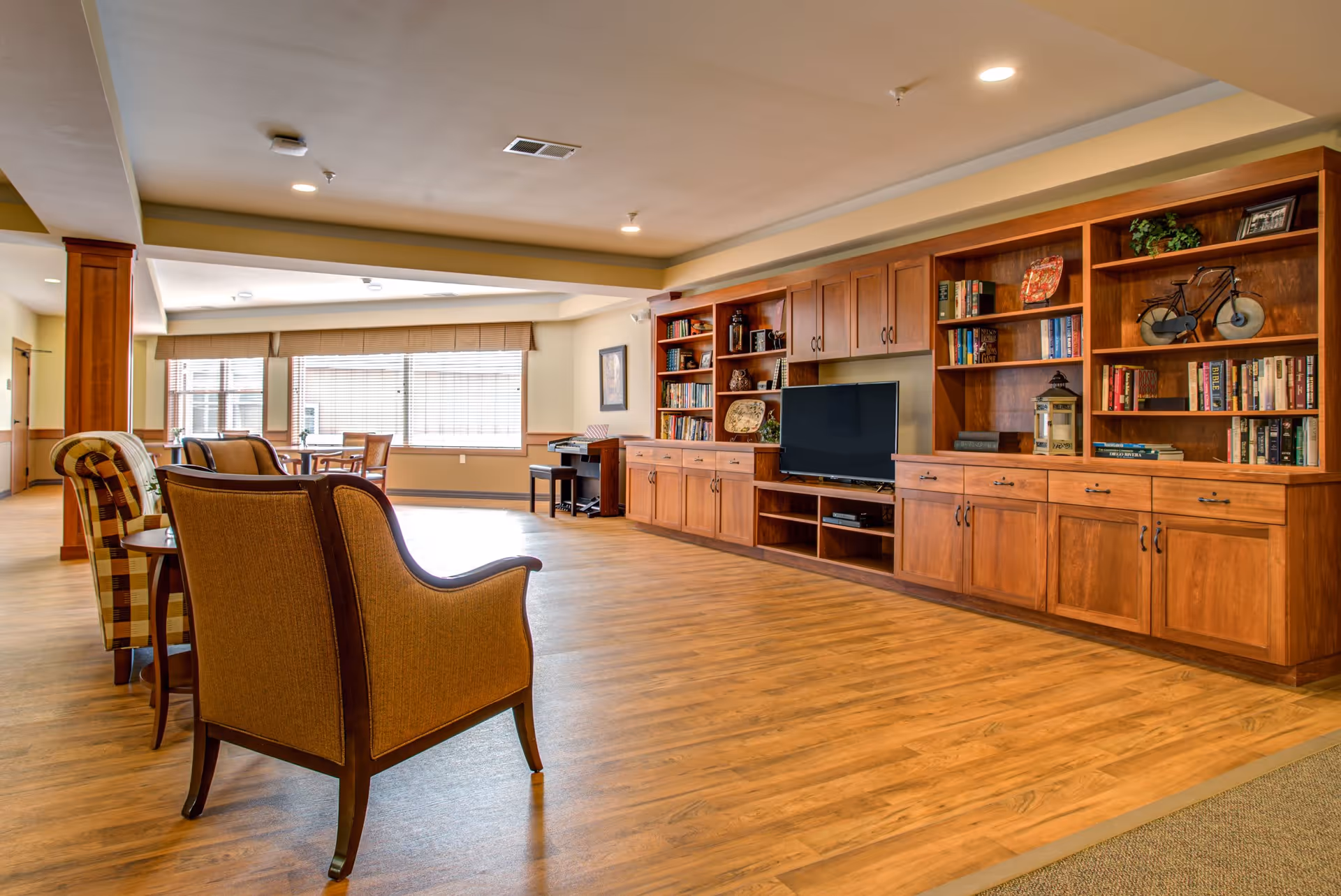 A spacious senior living common area with wooden flooring, comfortable armchairs, a large built-in wooden bookshelf filled with books and decorative items, and a flat-screen TV. Large windows allow natural light to brighten the room.
