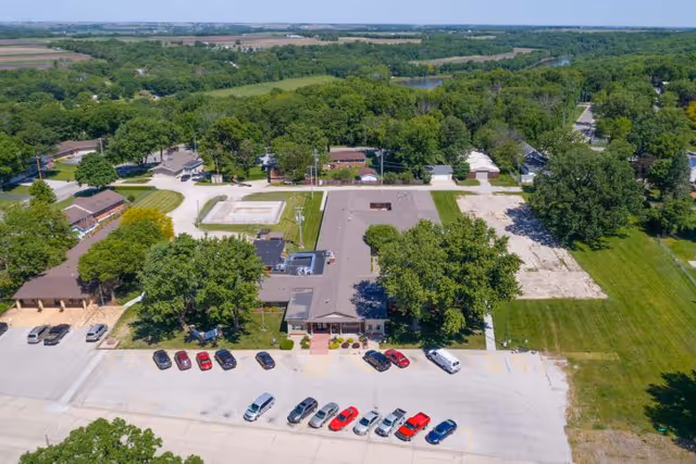 Aerial view of Fort Dodge Health and Rehabilitation facility surrounded by trees and greenery. The building has a flat roof and is situated near a parking lot with several cars parked. The surrounding area includes open grassy fields and a few other buildings in the distance.