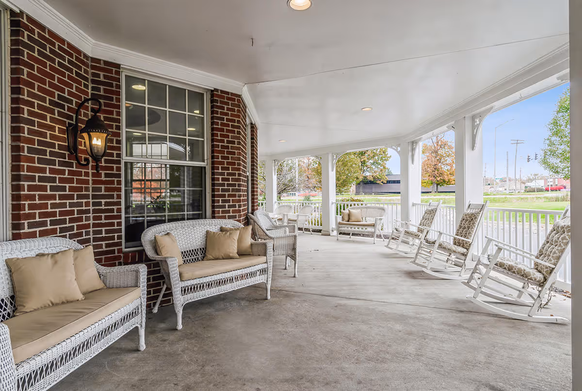 Covered front porch with white wicker sofas and rocking chairs along a brick wall overlooking a lawn.