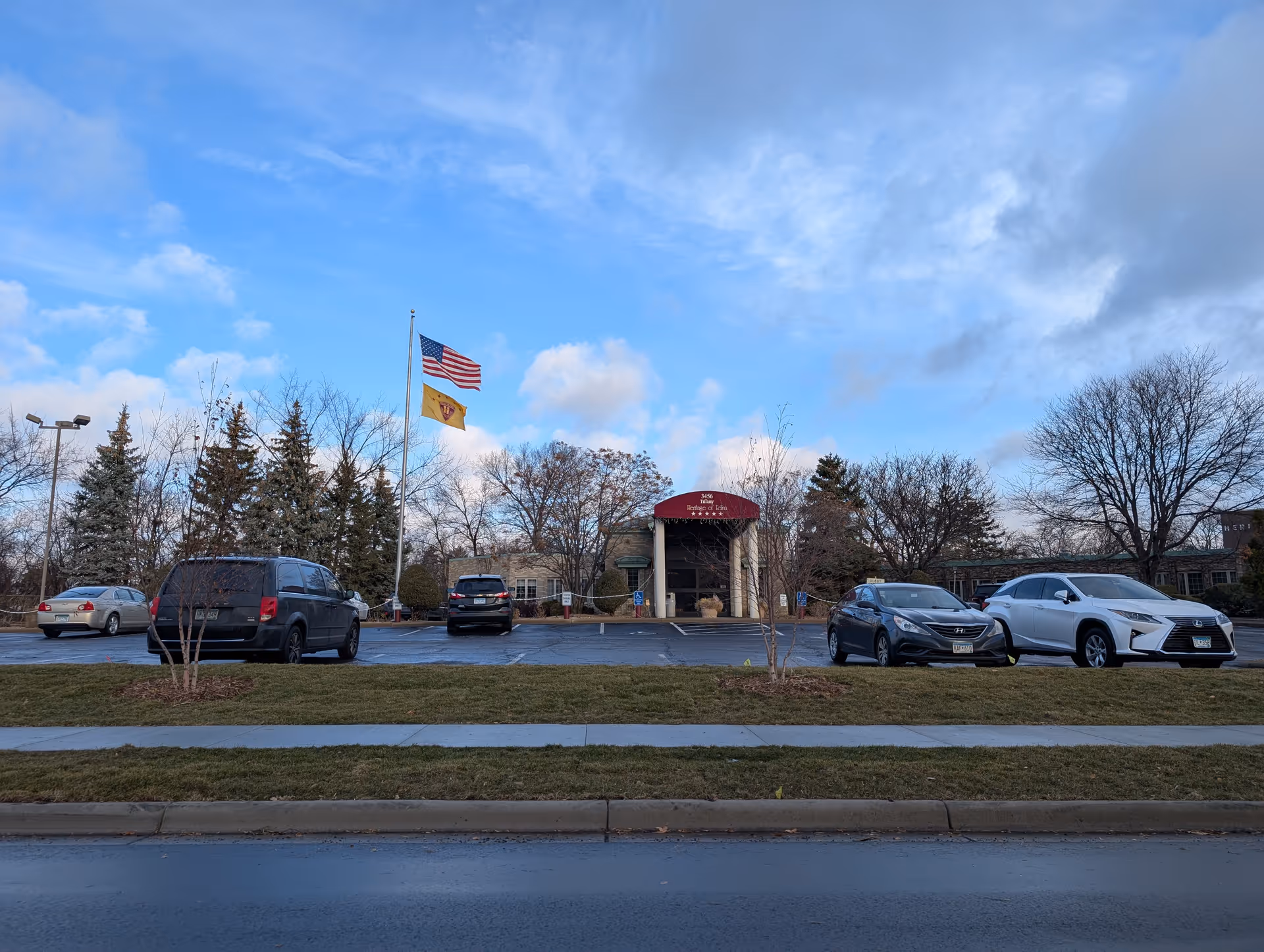 Front exterior of a senior living facility with a covered entrance, flagpoles, parked cars and a lawn under a blue sky.