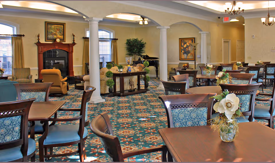 A spacious and well-lit common area with multiple wooden tables and chairs featuring blue patterned cushions. The room has a decorative carpet with a floral design, white columns, and arched openings. There is a fireplace with a wooden mantel, a grand piano, and framed artwork on the walls. Floral centerpieces are placed on the tables, and the room has a warm and inviting atmosphere.