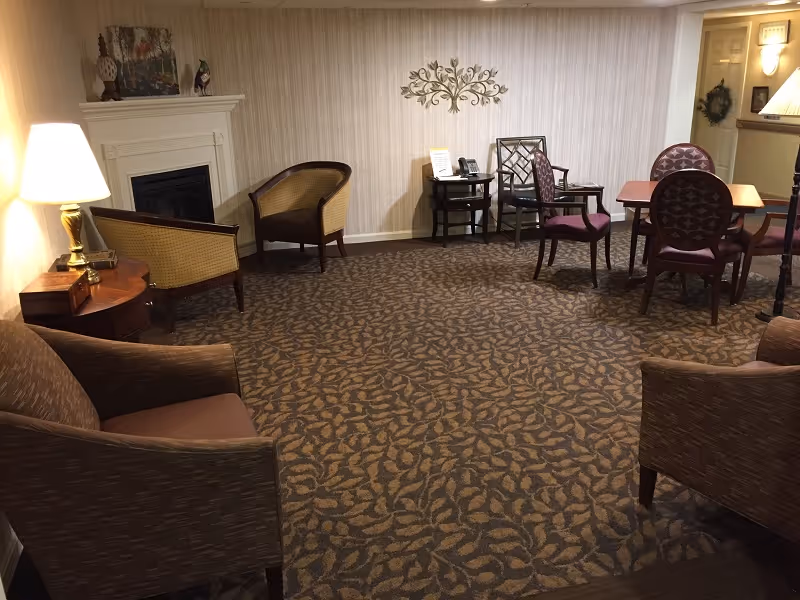 Carpeted communal lounge with upholstered chairs, small tables, a fireplace, and a dining table in the background.