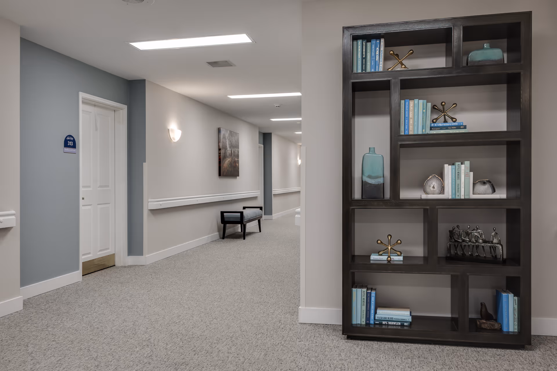A clean, well-lit hallway in a senior living facility with light gray walls and carpeted floor. On the left side, there is a white door labeled 313 and a bench along the wall. On the right side, there is a large dark wooden bookshelf with decorative items and books. The hallway extends into the distance with additional doors and wall-mounted lights.