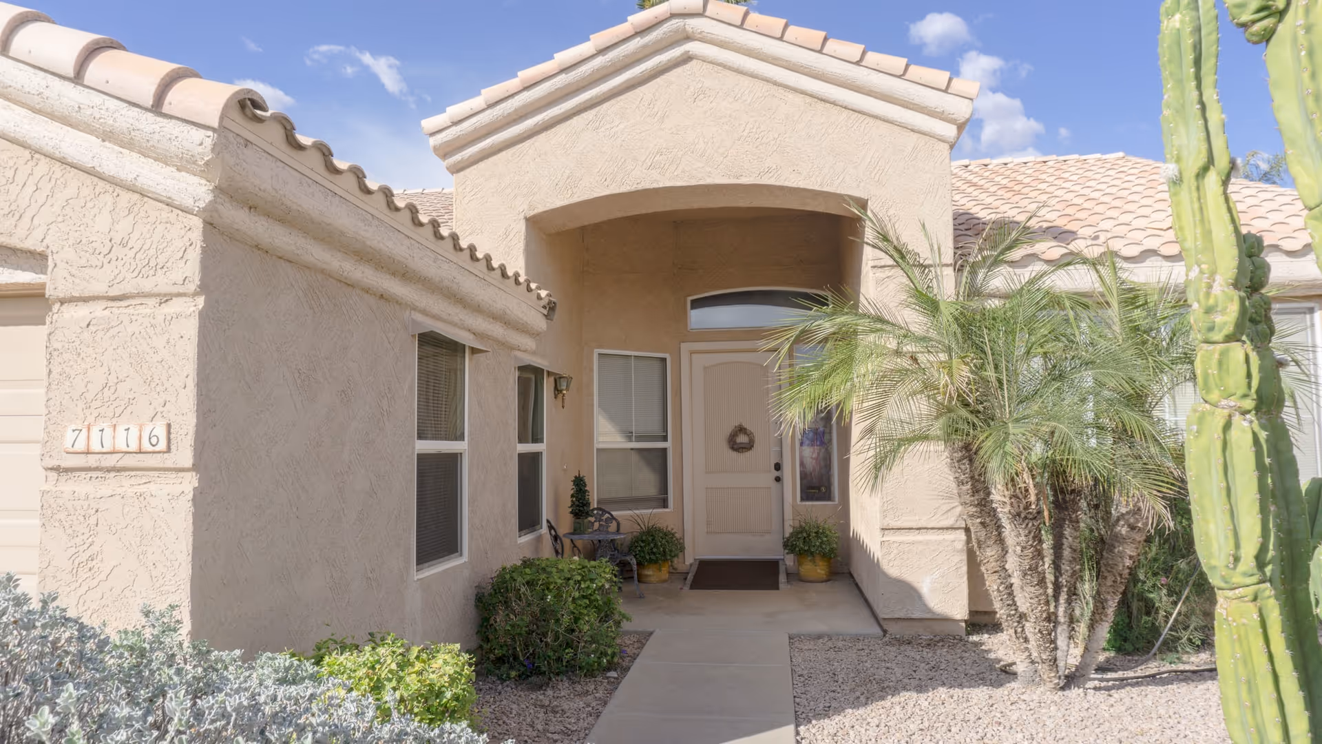 Front entrance of a beige stucco house with an arched porch, potted plants, palms and desert landscaping.