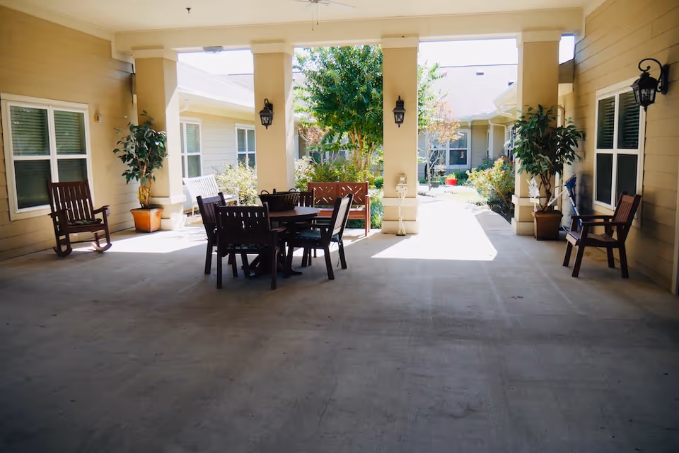 Covered outdoor courtyard with a round wooden table and chairs, rocking chairs, potted plants, and a view into a sunlit garden.
