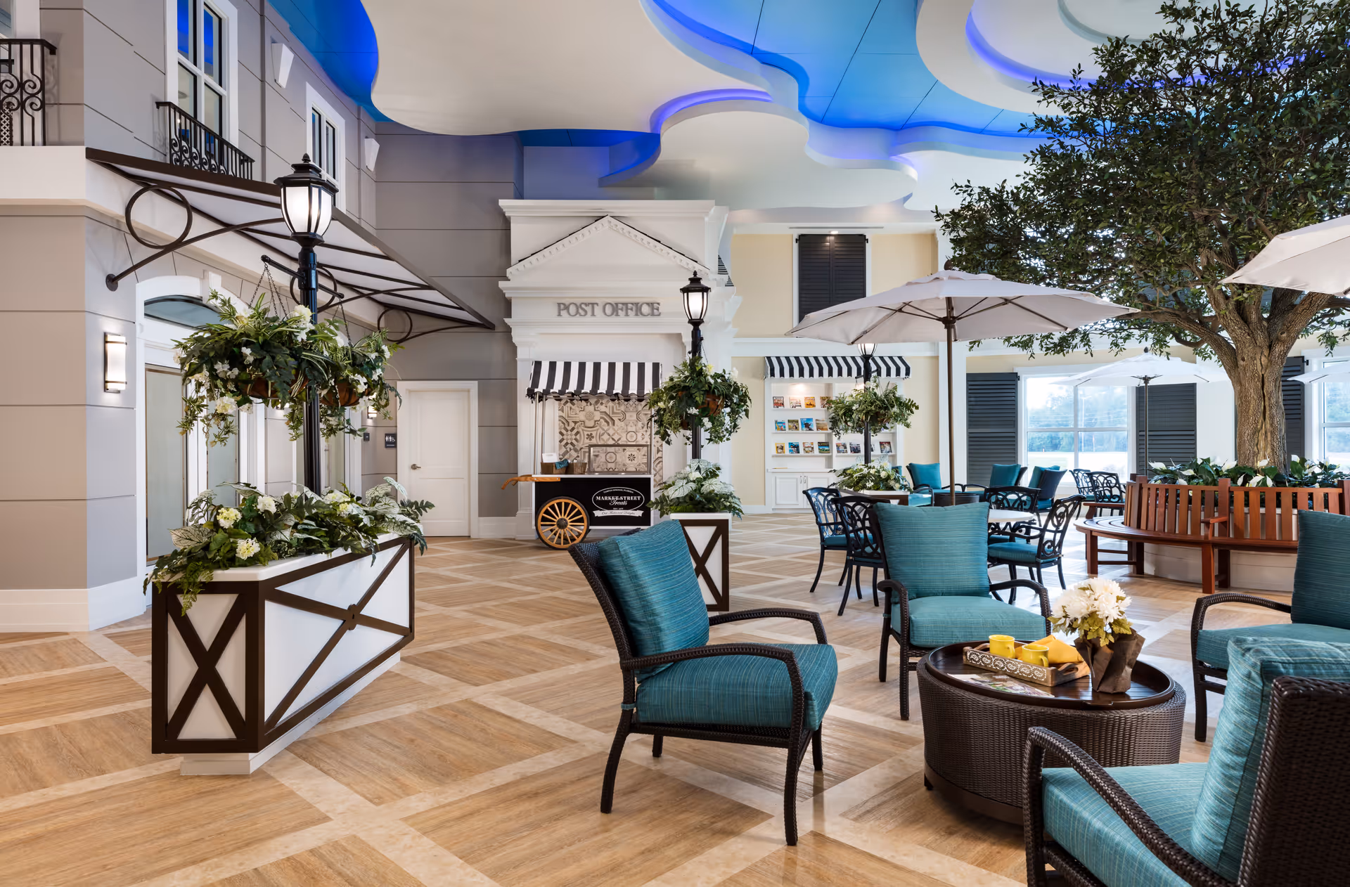 Indoor common area with comfortable teal cushioned chairs and round tables, some with umbrellas. There are decorative plants, a large tree with a circular bench around it, and a small post office facade with a striped awning. The ceiling has a modern design with blue lighting accents.