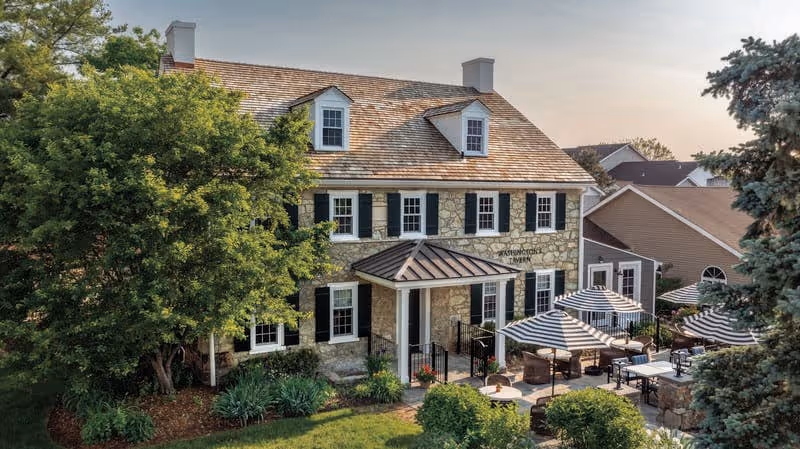 Stone building with a shingled roof and black shutters surrounded by trees and greenery. In front of the building, there is a patio area with tables and chairs shaded by striped umbrellas. The building has a sign that reads 'Washington's Tavern'.