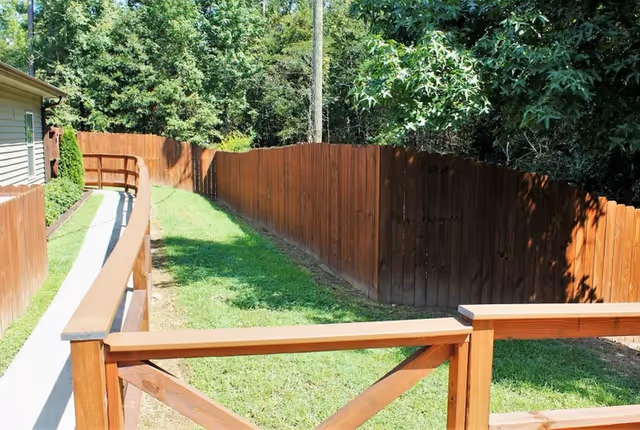 Grassy side yard with a wooden fence, ramped walkway and deck railing alongside a building with trees in the background.
