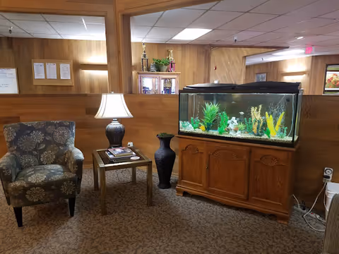 A cozy lounge area with an armchair, side table and lamp next to a large fish tank on a wooden cabinet in a wood-paneled room.
