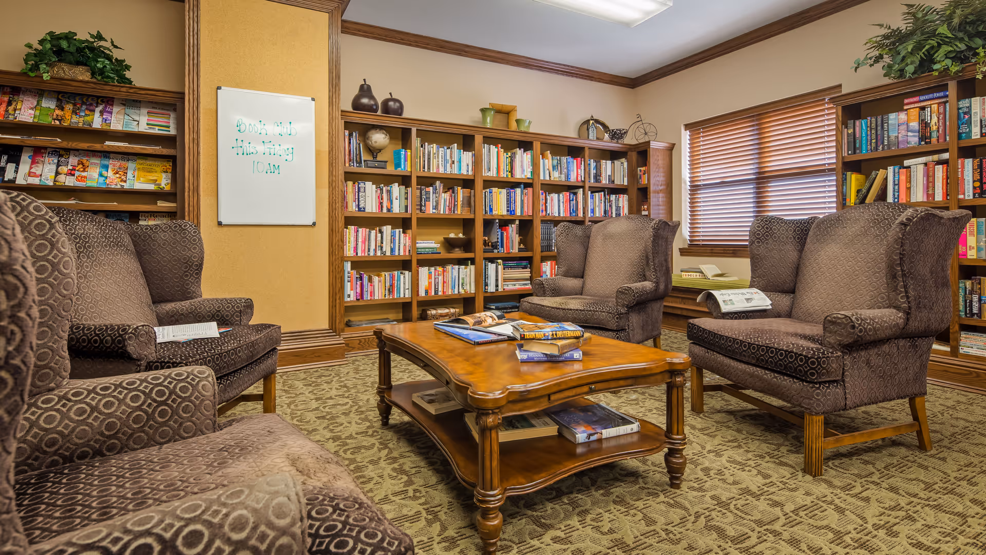 A cozy library room in a senior living facility with four patterned armchairs arranged around a wooden coffee table. The table holds several books and magazines. The room features bookshelves filled with books and magazines, a window with wooden blinds, and a carpeted floor with a patterned design. There is a whiteboard on the wall with a note about a book club meeting.