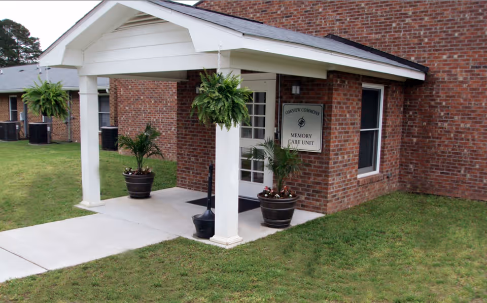 Entrance to the Oakview Commons Memory Care Unit at a senior living facility, featuring a white covered porch with hanging plants and potted plants on either side of the door, set against a red brick building with a grassy lawn.