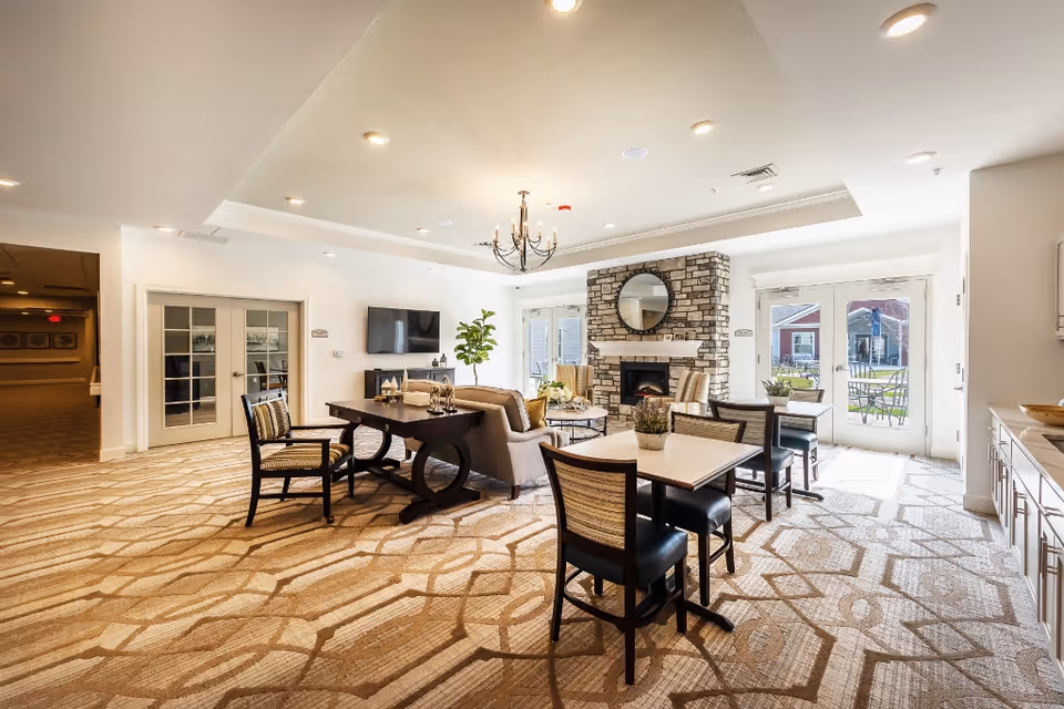 A bright and spacious common area with patterned carpet, multiple tables and chairs, a sofa, and a stone fireplace with a round mirror above it. There is a TV mounted on the wall, a chandelier hanging from the ceiling, and large glass doors leading outside to a patio with outdoor seating.