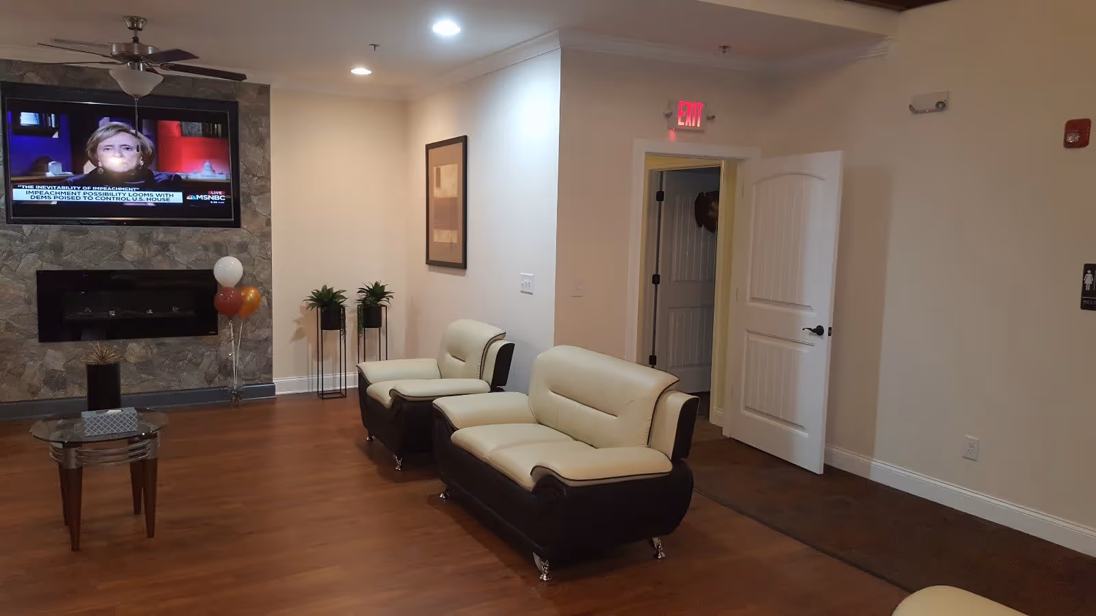A cozy senior living facility common area with two cream and brown leather armchairs and a matching loveseat on a wooden floor. A flat-screen TV is mounted on a stone wall above a modern fireplace. There are three balloons in white, orange, and gold near the fireplace, two potted plants on stands, a small glass coffee table with a tissue box, and an open door leading to another room. An exit sign is visible above the door.
