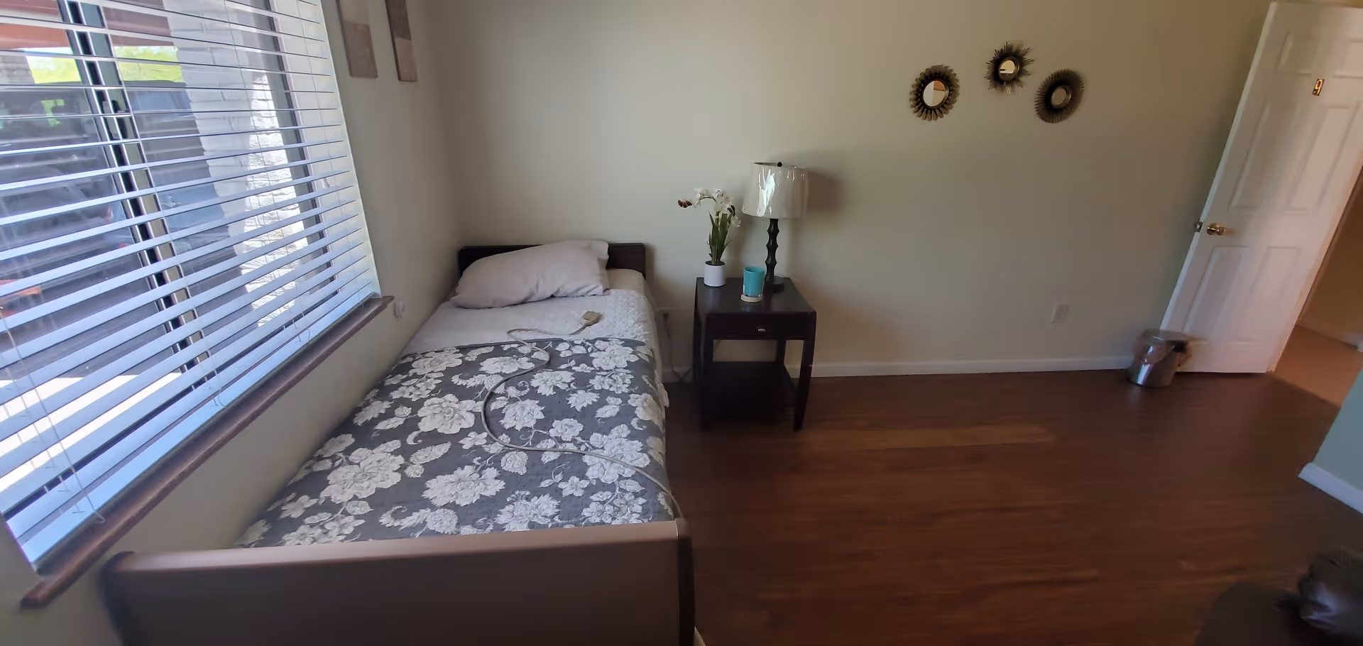 A small bedroom with a single bed covered in a floral patterned blanket and a pillow. Next to the bed is a dark wooden nightstand with a lamp, a small plant, and a blue cup. The room has wooden flooring, a window with blinds on the left wall, and three decorative round mirrors on the wall above the nightstand. A white door is open on the right side of the room, and a small trash can is visible near the door.