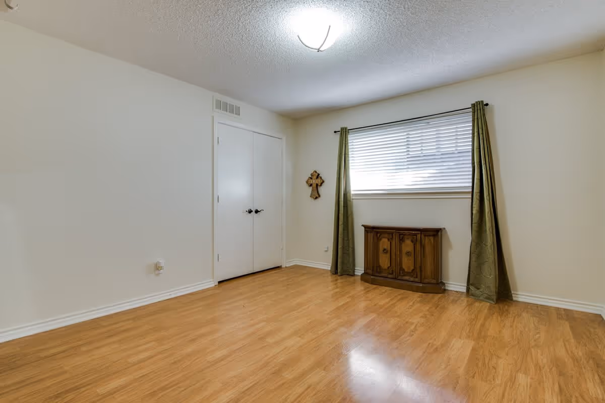 Empty room with light wood flooring, white walls, a window with green curtains and blinds, a small wooden cabinet beneath the window, a double white door closet, and a ceiling light fixture.