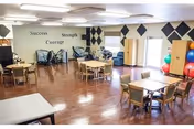 A spacious room with wooden flooring featuring several tables and chairs arranged for group activities or dining. The walls have motivational words such as 'Success', 'Strength', and 'Courage'. Exercise equipment and colorful therapy balls are visible near the windows, suggesting a multi-purpose activity or therapy room.