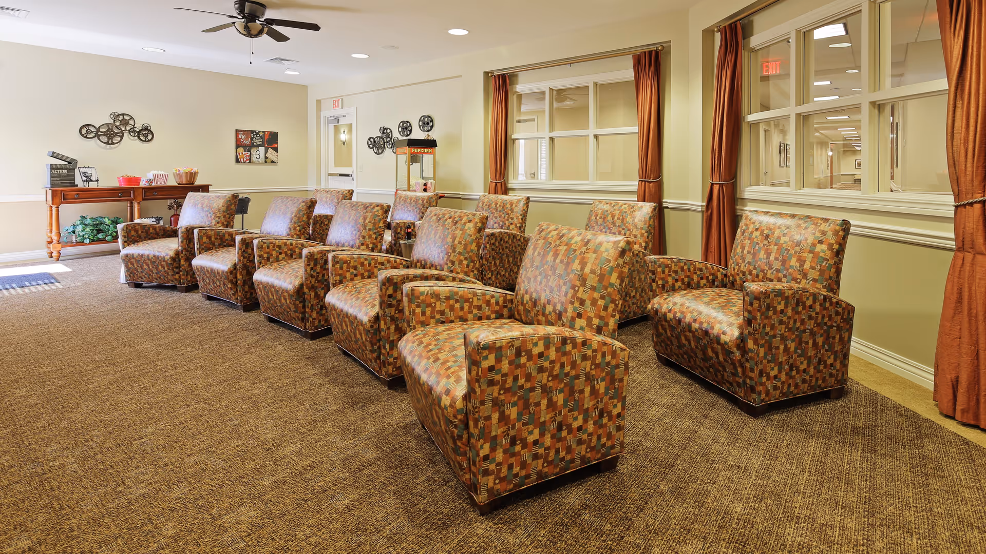 A cozy seating area with two rows of patterned armchairs arranged facing forward in a room with beige walls and carpeted floor. The room features large windows with rust-colored curtains, a ceiling fan, and a wooden table in the back holding popcorn supplies and decorative items.