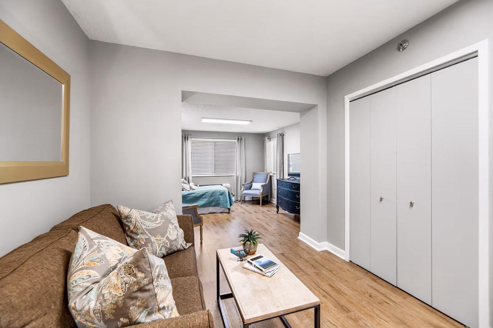 A cozy living area with a brown sofa adorned with patterned pillows, a small coffee table with books and a plant, and a large mirror on the wall. Beyond the living area is a bedroom with a bed covered in a teal blanket, a blue armchair, a dresser with a TV, and windows with blinds and curtains. The room has light gray walls and wood flooring.