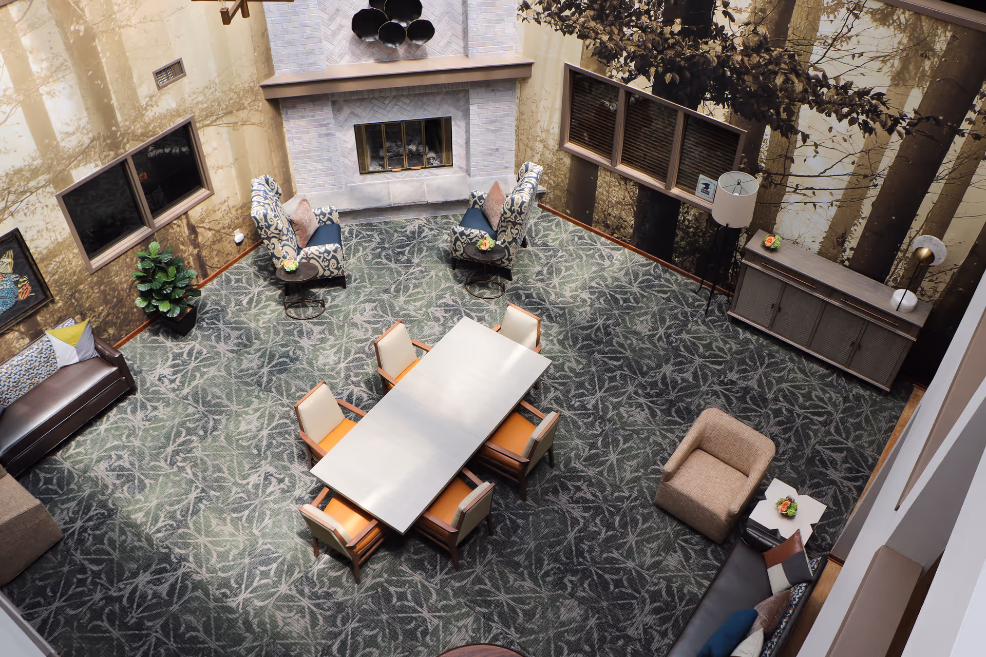 A cozy common area with a patterned carpet, a rectangular table surrounded by six chairs with orange cushions, two patterned armchairs near a fireplace, a beige armchair, and multiple sofas. The walls feature large nature-themed murals with tree images, and there are windows, a floor lamp, and a sideboard with decorative items.