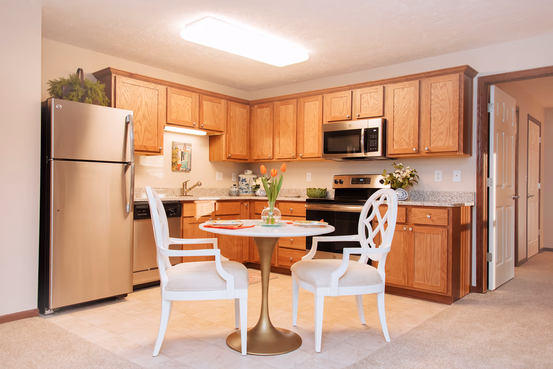 A bright kitchen area with wooden cabinets, stainless steel refrigerator, dishwasher, stove, and microwave. In the center, there is a small round table with a gold pedestal base and two white cushioned chairs. A vase with orange tulips is on the table. The kitchen floor is tiled, and the adjacent room has carpeted flooring.