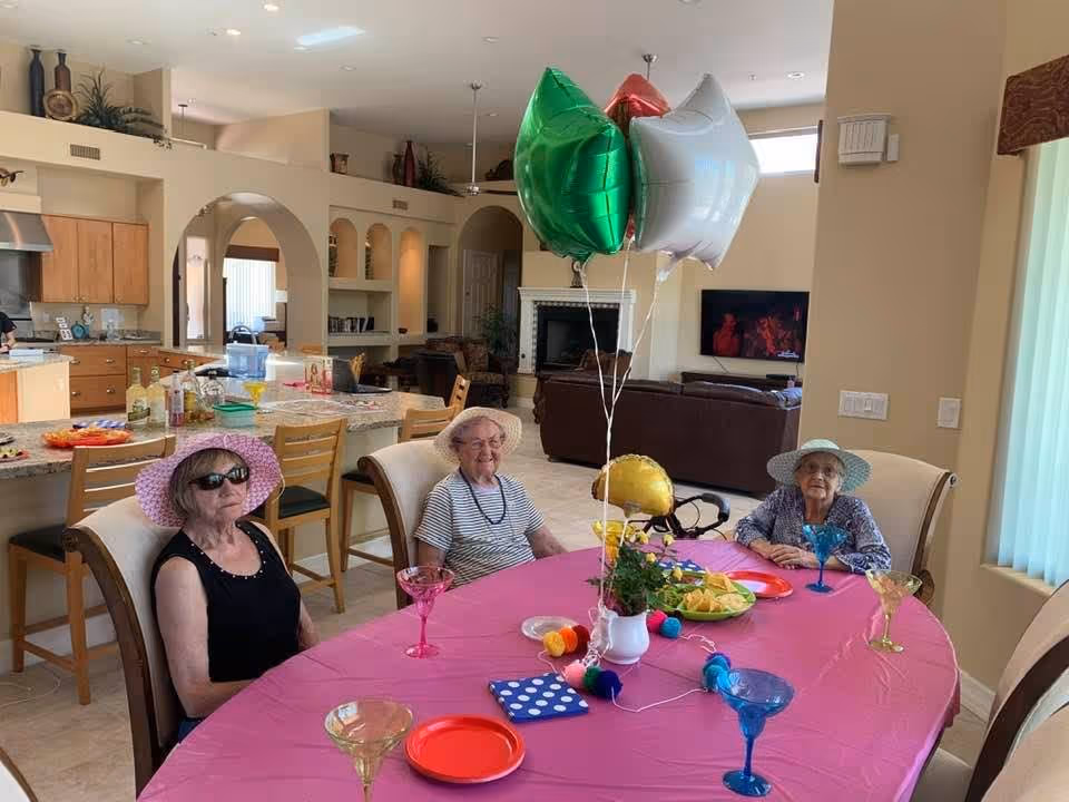 Three elderly women wearing sun hats sitting around a dining table covered with a pink tablecloth. The table has colorful plates, glasses, a small plant, and balloons in green, red, white, and gold. The background shows a spacious, well-lit kitchen and living area with wooden cabinets, a fireplace, and a television.