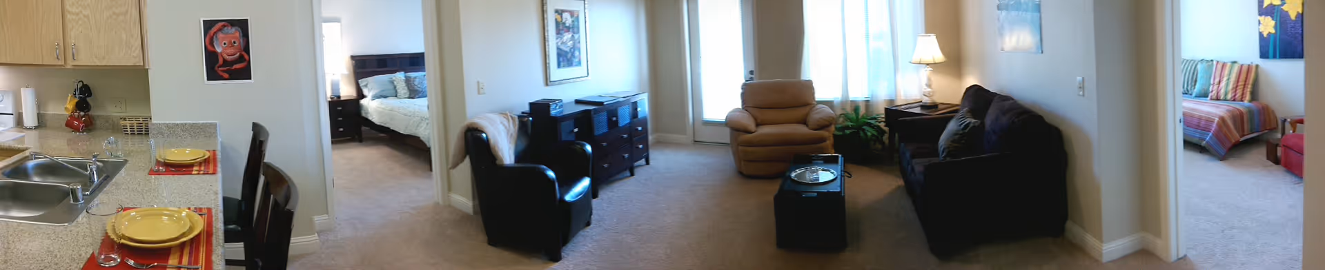 Panoramic view of an interior living space in a retirement community showing a kitchen area with a sink and two place settings on a counter, a living room with armchairs, a recliner, a coffee table, and lamps, and two adjacent bedrooms visible through doorways.