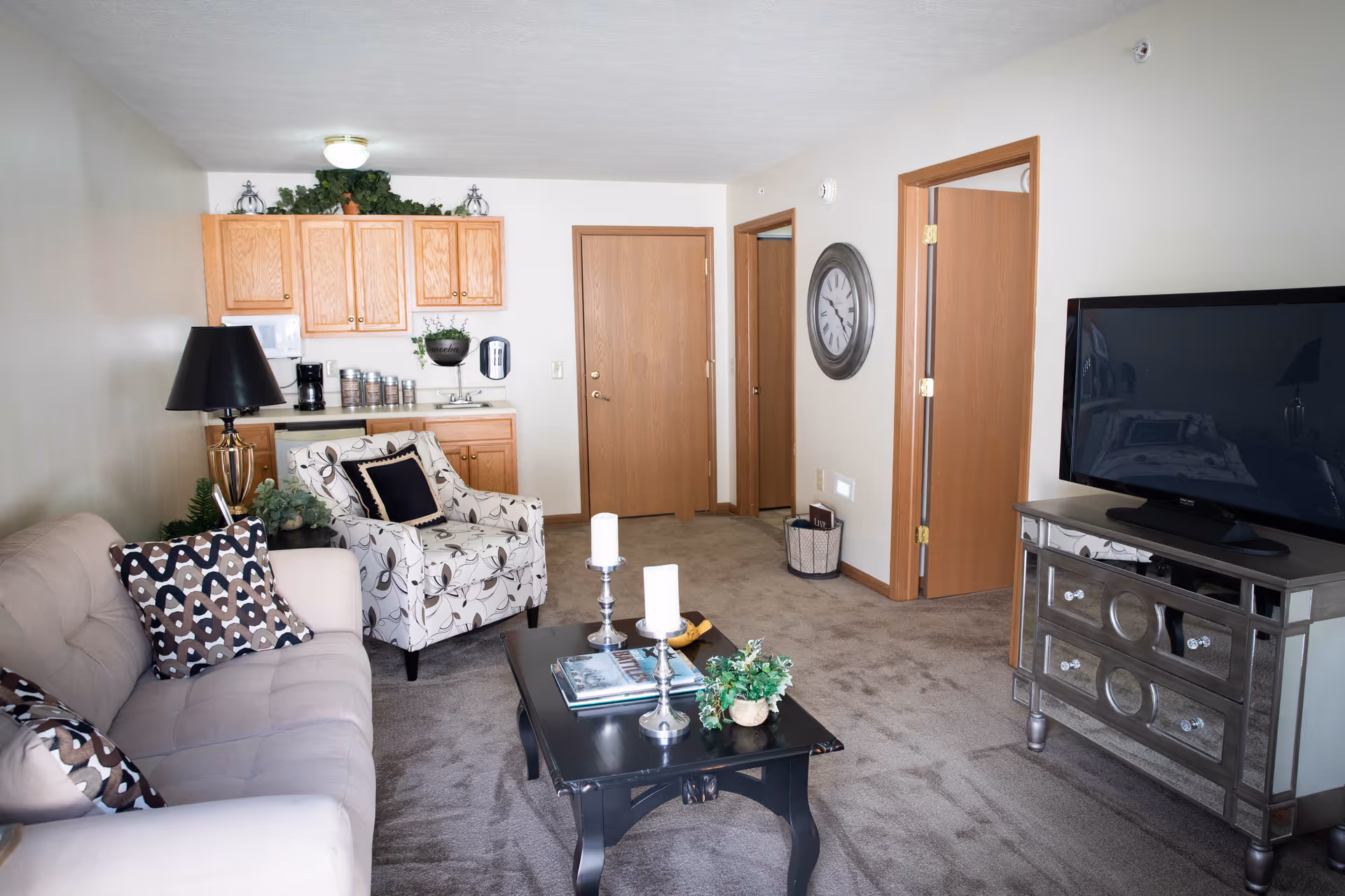 A cozy living room area in an assisted living facility featuring a beige sofa with patterned pillows, a floral armchair, a black coffee table with candles and a small plant, a mirrored TV stand with a flat-screen TV, and a kitchenette with wooden cabinets and a coffee maker in the background. The room has beige carpet and light-colored walls with wooden doors and trim.