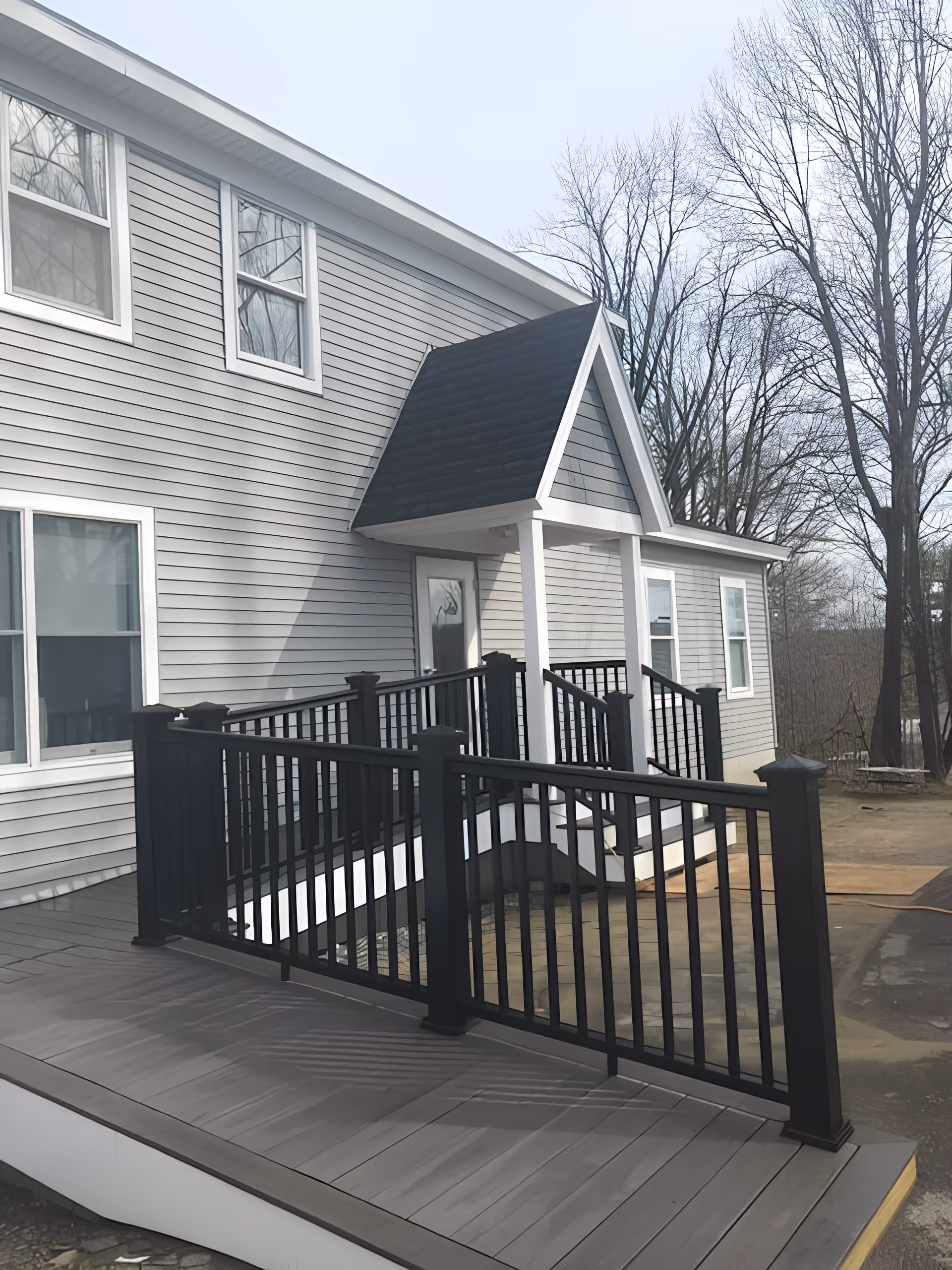 Exterior view of a gray two-story building with white trim and a black shingled small roof over the entrance. There is a black metal railing along a ramp leading up to the door. Leafless trees are visible in the background under a clear sky.