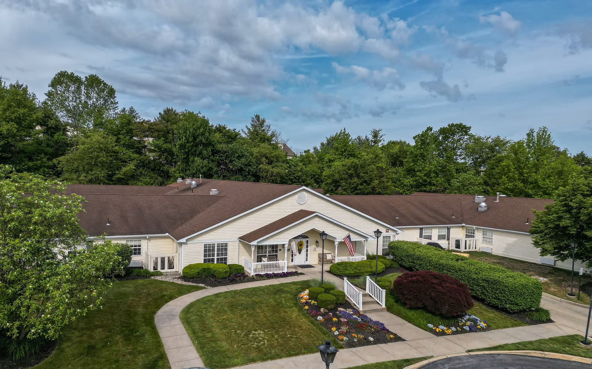Exterior view of a single-story building with a brown roof and cream-colored siding, surrounded by well-maintained landscaping including bushes, trees, and flower beds. A curved sidewalk leads to the entrance, which has a small covered porch with an American flag displayed. The building is set against a backdrop of tall green trees under a partly cloudy sky.