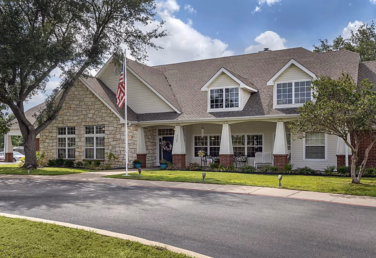 Exterior view of a senior living facility building with a stone and siding facade, a covered porch with seating, an American flag on a flagpole, and trees and landscaping around the entrance under a partly cloudy sky.