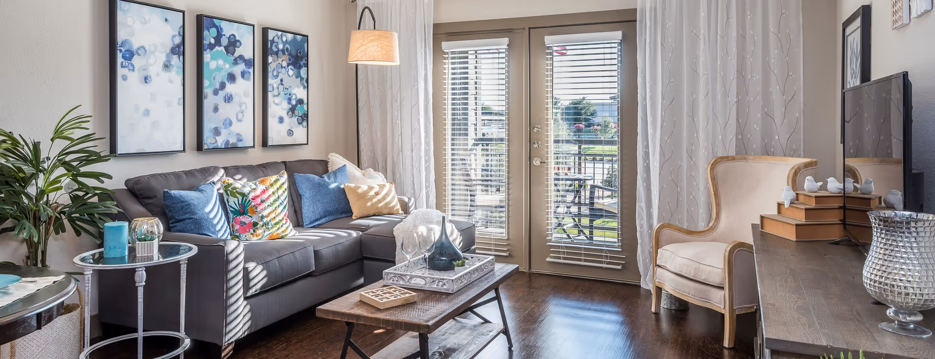 A bright and cozy living room with a gray sectional sofa adorned with colorful pillows, a wooden coffee table with decorative items, a beige armchair, a flat-screen TV on a wooden console, and glass doors with blinds leading to an outdoor patio. The room features three abstract blue and white paintings on the wall and a floor lamp beside the sofa.