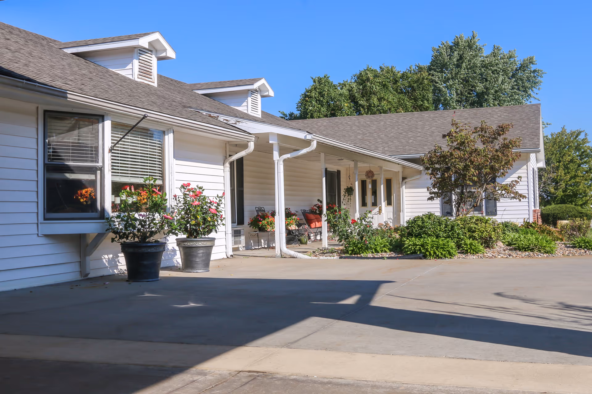 Exterior view of a white single-story building with a gray roof, featuring a covered porch with columns, potted plants, and greenery along the walkway under a clear blue sky.